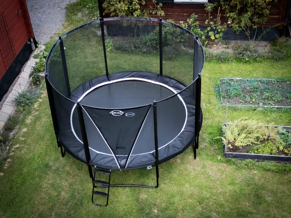 Studsmatta monterad med skyddsnät, kantskydd och stege i trädgårdsmiljö.